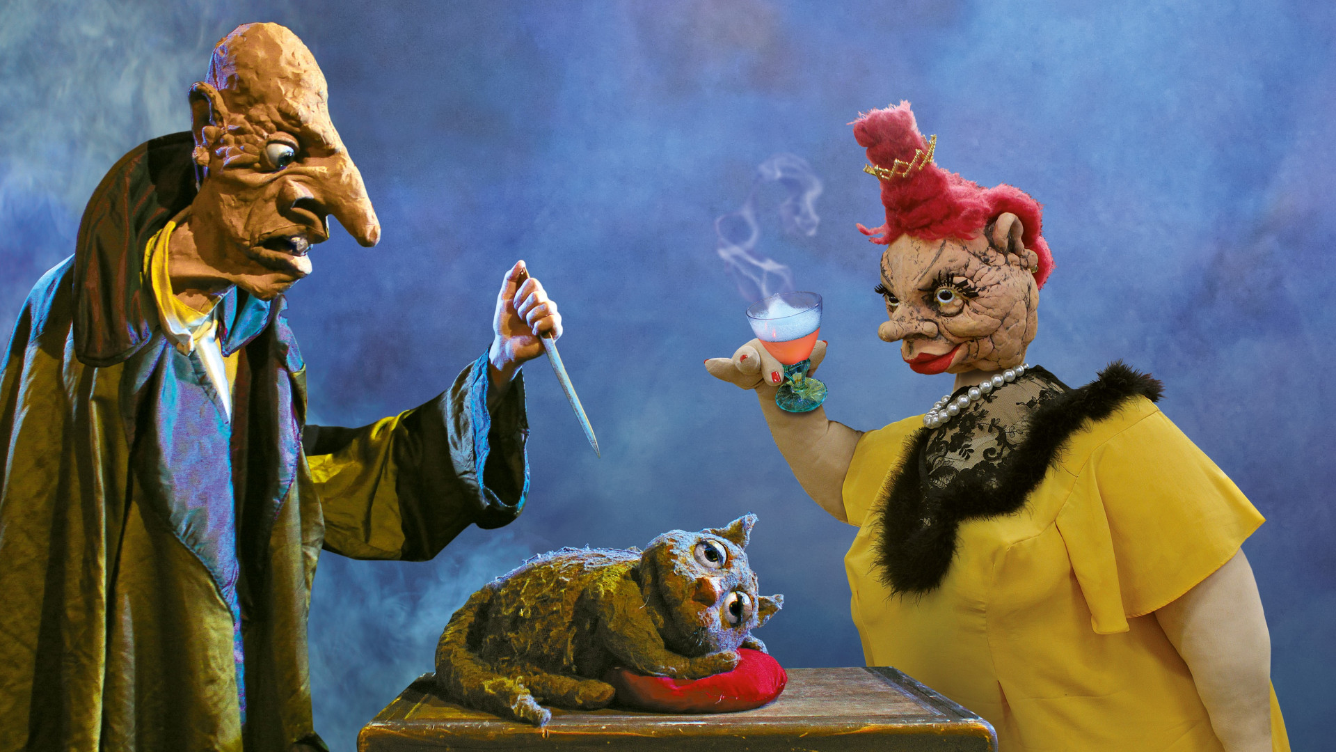 The image shows two puppet-like figures in a surreal and eerie scene. On the left, a bald, hunched-over character with exaggerated facial features and a long, crooked nose holds a knife over a small creature resembling a worn, distressed cat laying on a table. On the right, a larger, female puppet with bright pink hair styled in a messy updo, wearing a yellow dress and black fur trim, holds a smoking cocktail glass. The background is a smoky, ominous blue haze, giving the scene a dark, theatrical, and otherworldly feel.