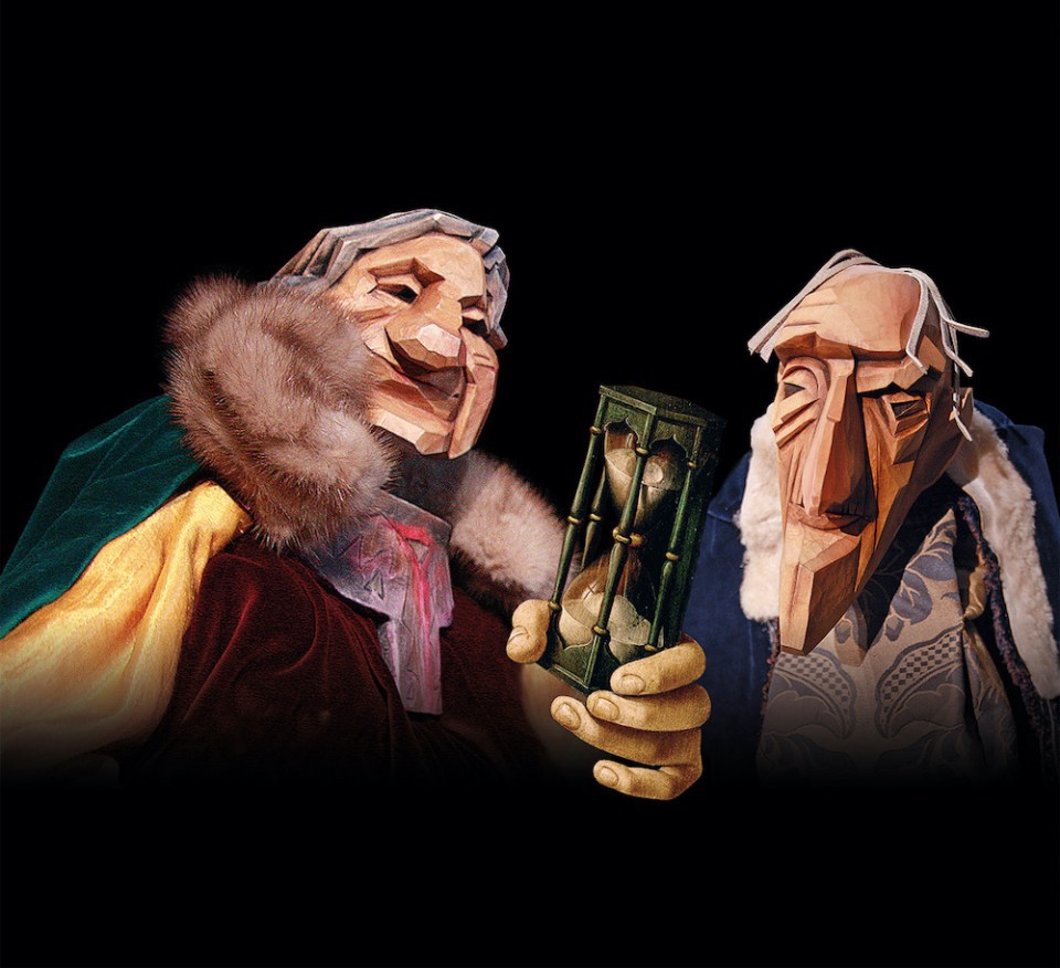 The image depicts two puppet figures with intricately carved wooden faces, standing against a solid black background. The character on the left is dressed in a fur-lined, regal robe, holding an hourglass, while the figure on the right wears a patterned outfit with a somber expression. The hourglass, a symbol of time passing, is central to the scene, and both characters appear elderly and reflective, adding a contemplative, philosophical tone to the image.