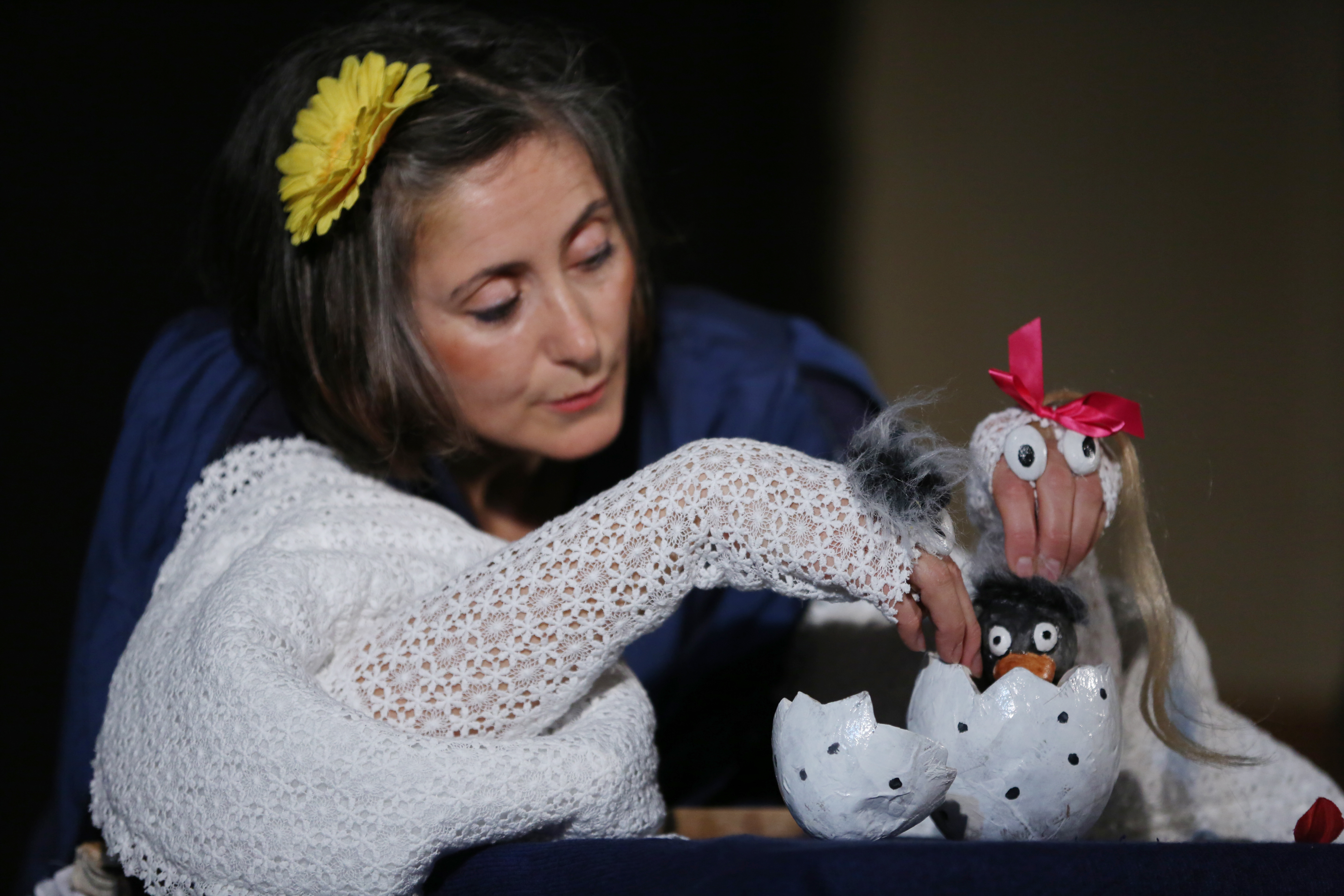 Eine Frau mit gelber Blume im Haar spielt mit Puppen: Eine kleine schwarze Figur schaut aus einer aufgebrochenen Eierschale, daneben eine Handpuppe mit Schleife. Der Hintergrund ist dunkel.