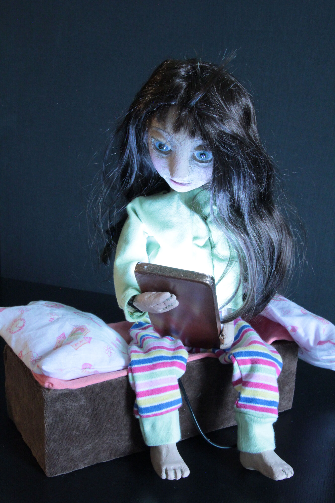 Eine Figur mit langen, dunklen Haaren sitzt auf einer kleinen Bank mit Kissen, trägt bunte Schlafanzughosen und hält ein Tablet in den Händen, vor dunklem Hintergrund.