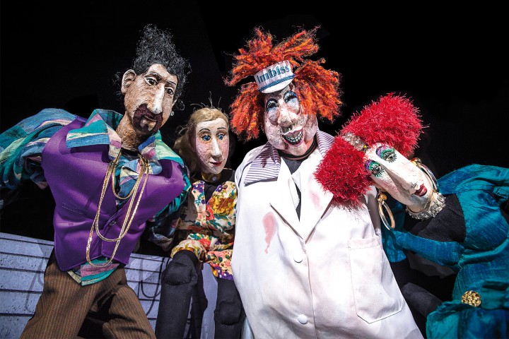 Vier bunte Marionetten in auff&auml;lligen Kost&uuml;men und mit unterschiedlichen Frisuren, darunter eine Figur mit rotem Haar und Imbiss-M&uuml;tze, stehen dicht beieinander vor schwarzem Hintergrund.