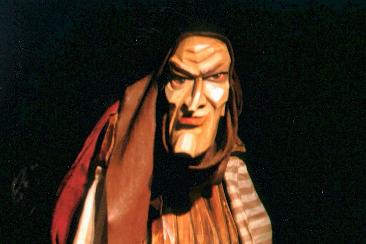 Holzfigur mit markanten Gesichtsz&uuml;gen in mittelalterlicher Kleidung sitzt auf einer Bank, dramatisch beleuchtet.