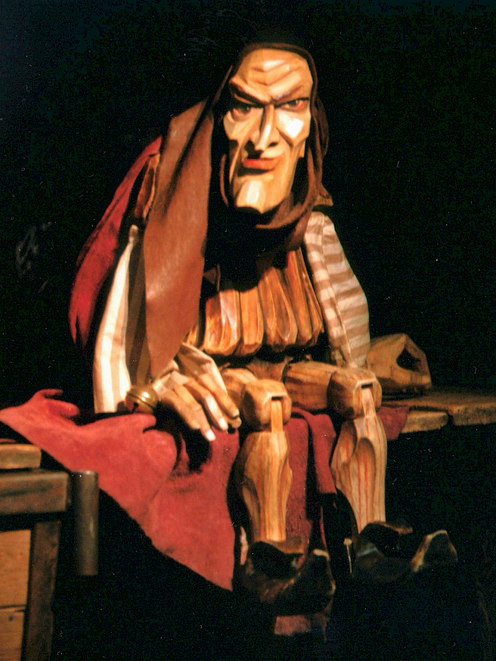Holzfigur mit markanten Gesichtszügen in mittelalterlicher Kleidung sitzt auf einer Bank, dramatisch beleuchtet.