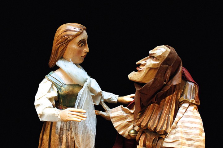 Holzfigur mit markanten Gesichtsz&uuml;gen in mittelalterlicher Kleidung sitzt auf einer Bank, dramatisch beleuchtet.