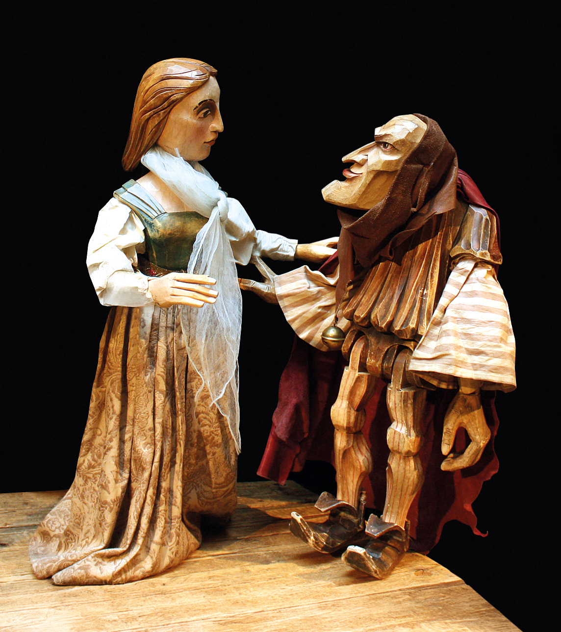 Holzfigur mit markanten Gesichtszügen in mittelalterlicher Kleidung sitzt auf einer Bank, dramatisch beleuchtet.