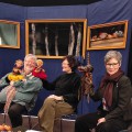 Drei Personen sitzen vor einem Puppentheater und halten verschiedene Hand- und Stabpuppen. Ein Tisch mit weiteren Puppen und Requisiten steht vor ihnen.