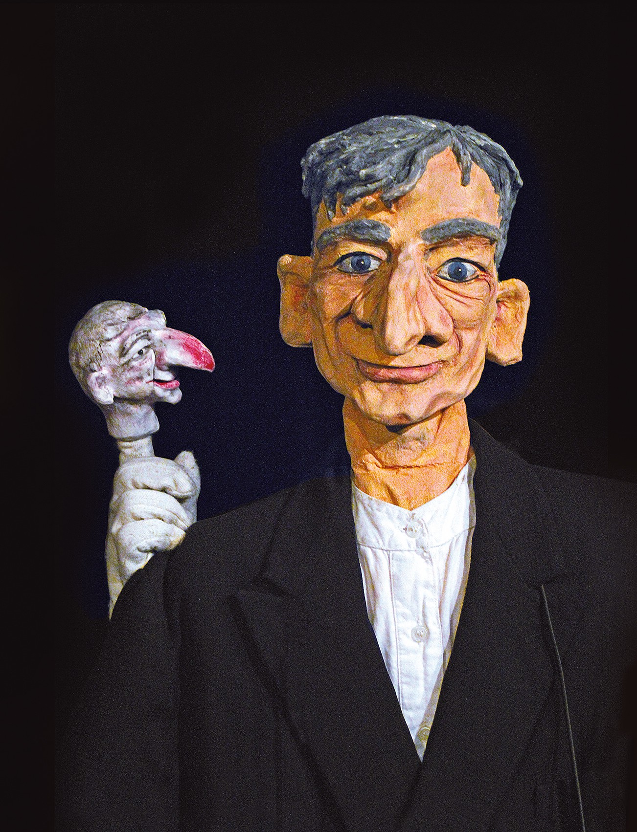 Ringelnatz Puppen.etc Eine Marionette mit markanten Gesichtszügen und kurzen grauen Haaren trägt einen schwarzen Anzug. Daneben eine kleinere, weiße Figur mit spitzer Nase, die in ihre Richtung blickt, vor schwarzem Hintergrund.