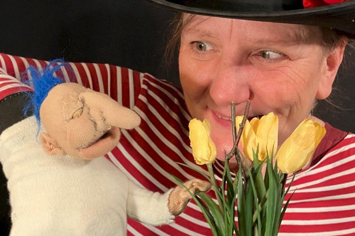 Frau mit schwarzem Hut und roter Blume hält eine Handpuppe, die auf gelbe Tulpen zeigt.