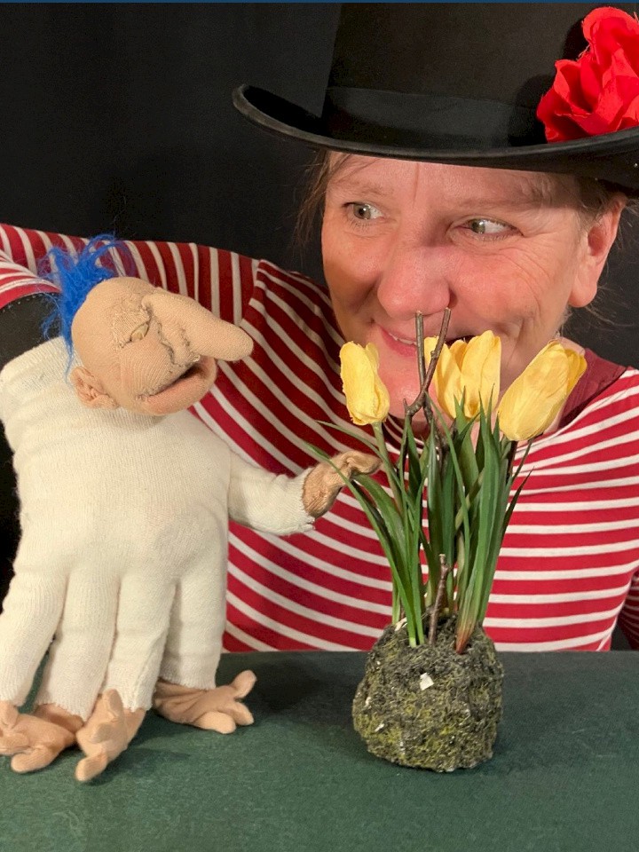 Frau mit schwarzem Hut und roter Blume hält eine Handpuppe, die auf gelbe Tulpen zeigt.