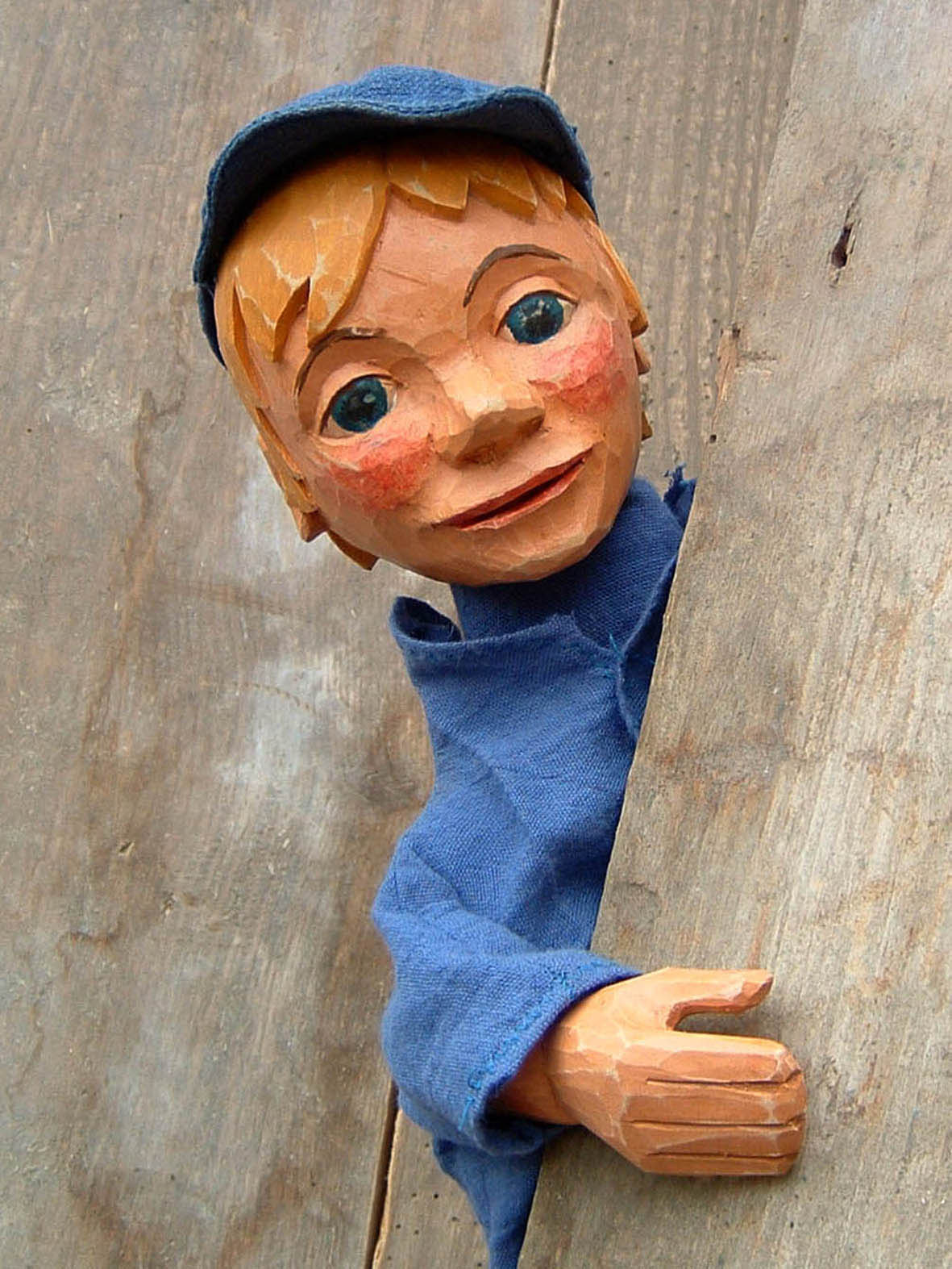 Presefoto Michel aus Lönneberga - Theater Blaues Haus Holzfigur eines Jungen mit blonden Haaren, blauen Augen und rotem Gesichtsausdruck, in blauer Kleidung und Mütze, schaut lächelnd hinter einem Holzbalken hervor.