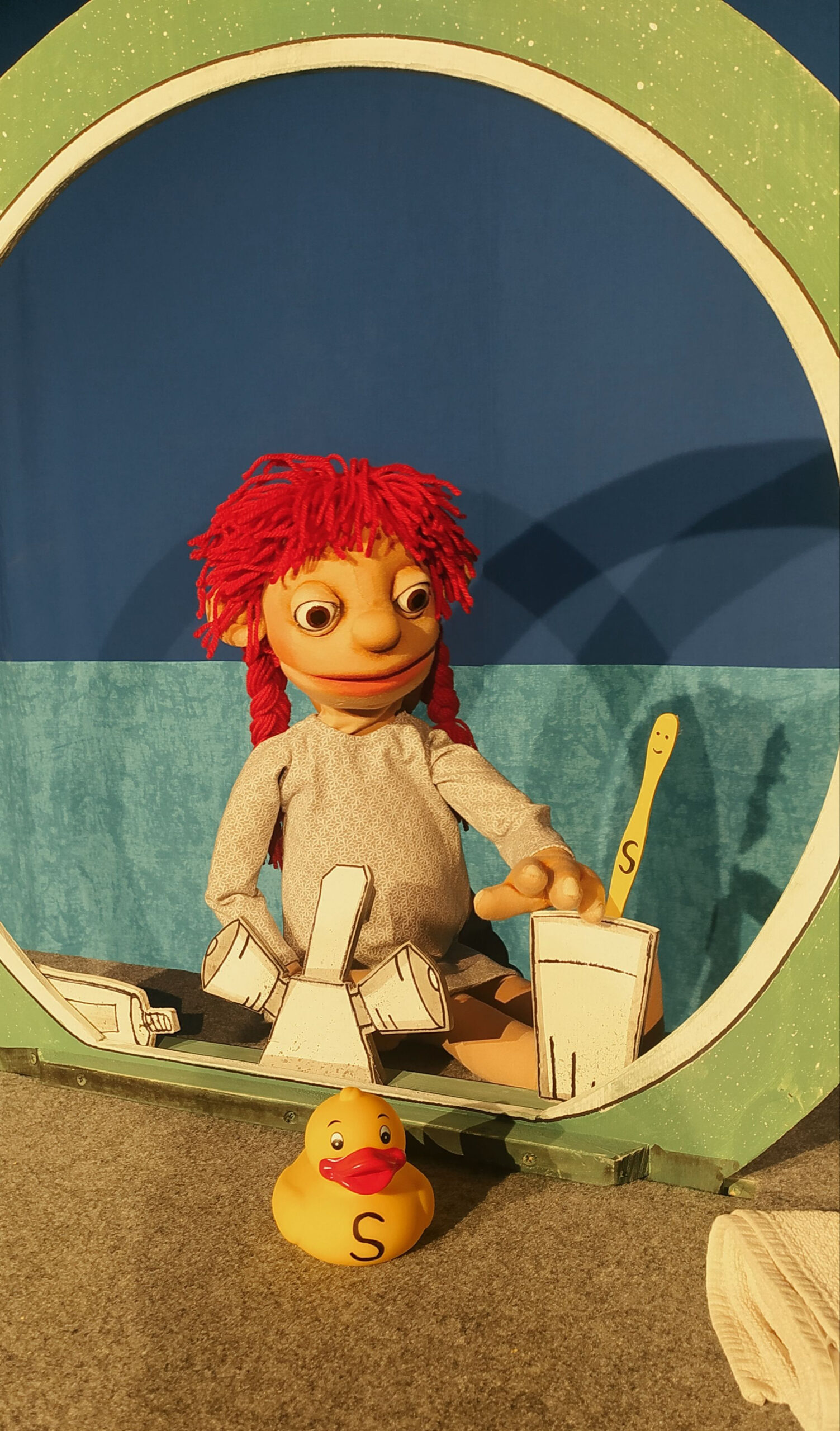 Sammy - Die ungewöhnliche Reise einer Zahnbürste Eine Marionette mit roten Zöpfen sitzt in einem stilisierten Boot vor blauem Hintergrund. Vor ihr liegt eine gelbe Badeente mit dem Buchstaben