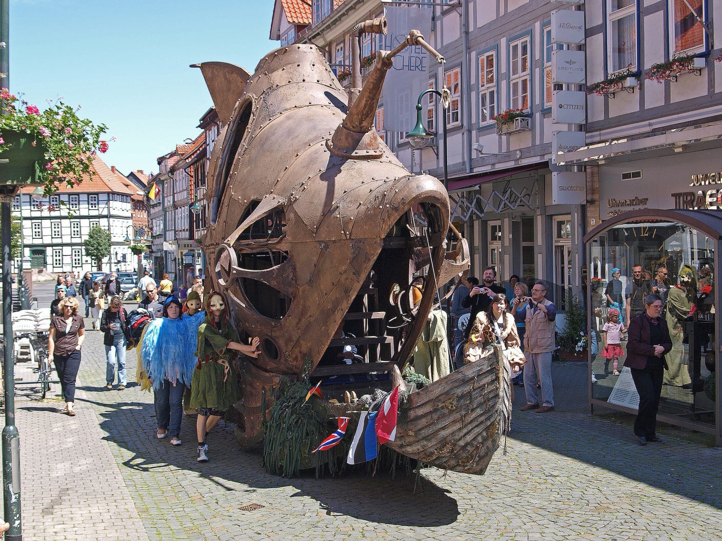 Eine belebte Altstadtstraße mit historischen Fachwerkhäusern, in der eine große, kunstvolle Metallkonstruktion in Form eines fischähnlichen, dampf-punkartigen Fahrzeugs oder Schiffs durch die Menge bewegt wird. Menschen in fantasievollen Kostümen mit Masken, Federn und bunten Gewändern begleiten das Gefährt. Zahlreiche Passanten stehen am Straßenrand und beobachten das Spektakel, einige machen Fotos.