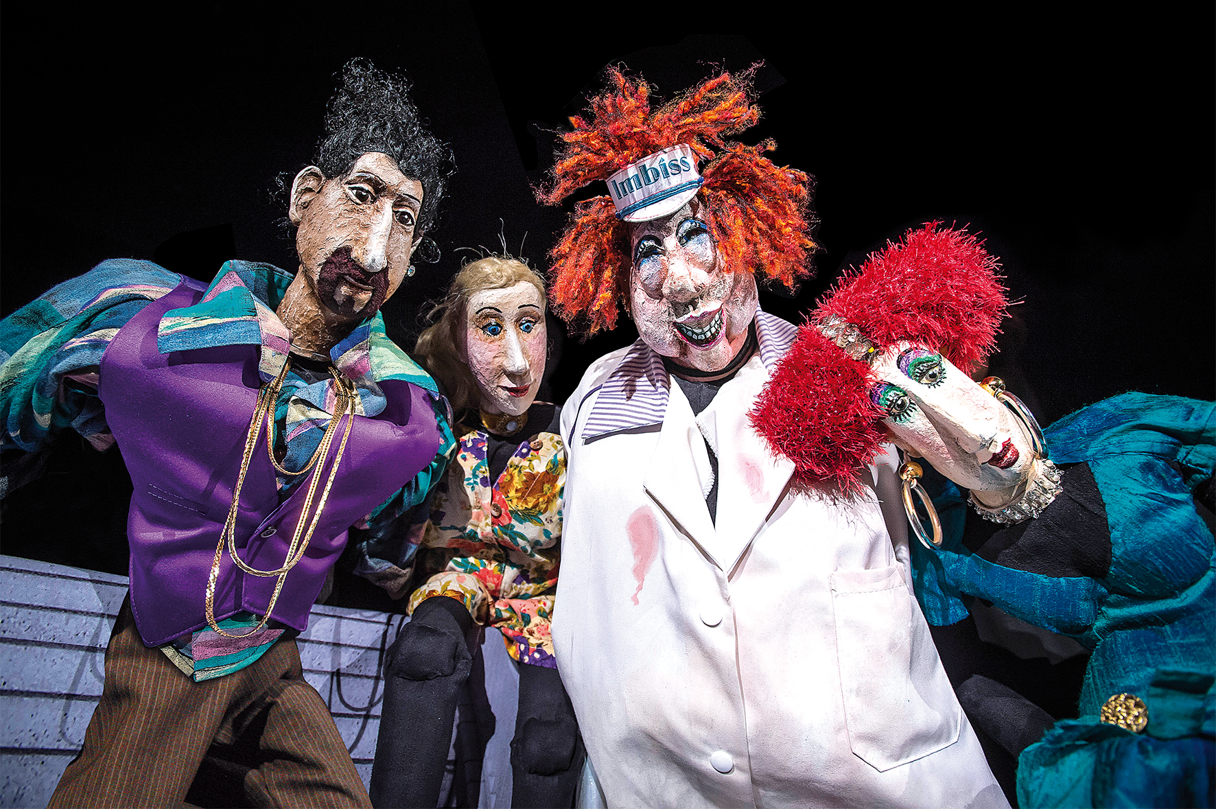 Vier bunte, theatralische Puppen mit übertriebenen Gesichtszügen und extravaganten Outfits vor dunklem Hintergrund.