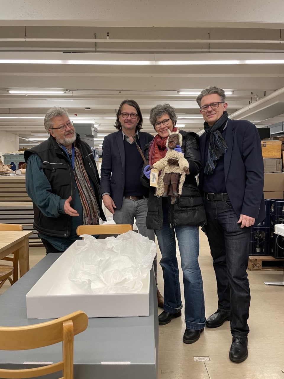 Vier Personen in einem Archivraum, eine davon hält eine Puppe, die einen Teufel darstellst, davor liegt eine geöffnete Archivbox auf dem Tisch.