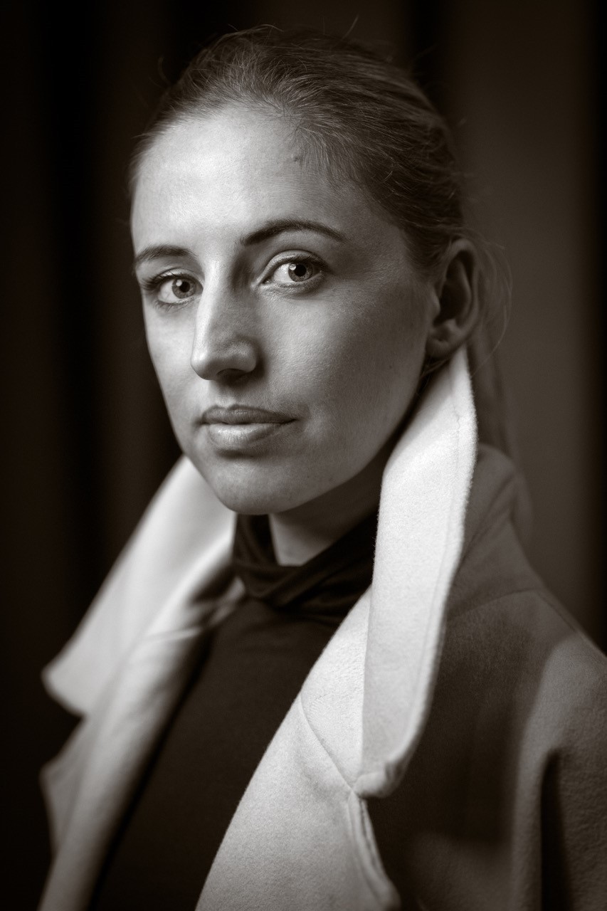 Schwarz-Weiß-Porträt einer Frau mit ernstem Blick, dezentem Make-up und zurückgebundenem Haar. Sie trägt einen hohen Kragen und einen eleganten Mantel, der ihre Schultern umrahmt.