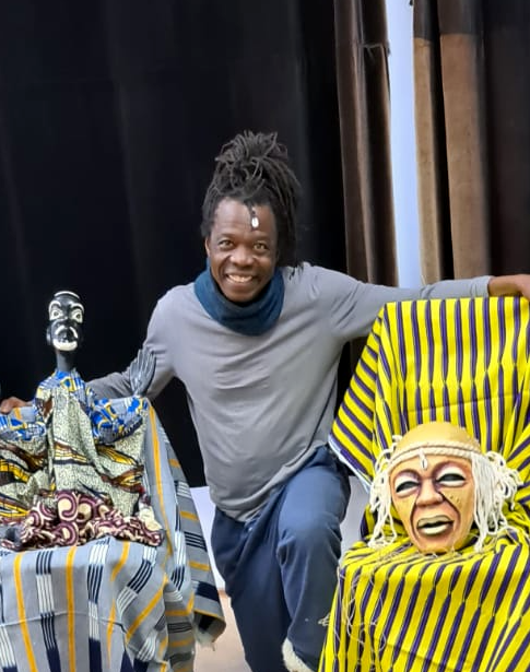 Ein lächelnder Mann mit Dreadlocks und grauem Shirt posiert zwischen zwei kunstvollen afrikanischen Puppen, die in bunte Stoffe gehüllt sind. Der Hintergrund besteht aus dunklen Vorhängen.