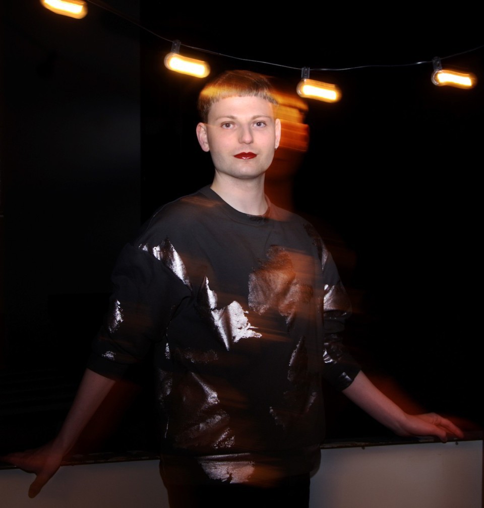 Porträt einer Person mit hellem Teint, rotem Lippenstift und modernem Kurzhaarschnitt, gekleidet in ein dunkles, glänzendes Oberteil. Der Hintergrund ist schwarz mit warmen Lichterketten.