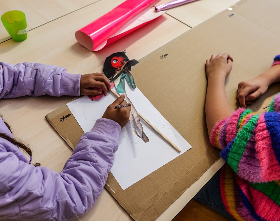 Das Bild zeigt zwei Kinder, die an einem Tisch sitzen und an einem Bastelprojekt arbeiten. Das Kind links trägt eine lila gesteppte Jacke und arbeitet mit einem Stift an einer Figur, die auf weißem Papier liegt. Auf dem Tisch befinden sich außerdem ein großes Stück Karton sowie rote Bastelmaterialien. Das Kind rechts trägt einen farbenfrohen Pullover mit bunten Streifen in Pink, Blau, Grün und Gelb. Beide Kinder sind mit dem Basteln beschäftigt und die Materialien deuten auf ein kreatives Projekt hin.