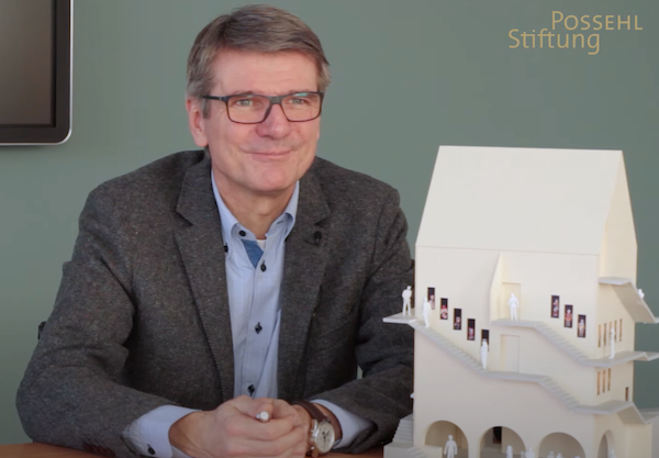 Ein Mann sitzt an einem Tisch und präsentiert ein Architekturmodell eines historischen Gebäudes mit Treppen und kleinen Figuren, die die Nutzung des Hauses andeuten.