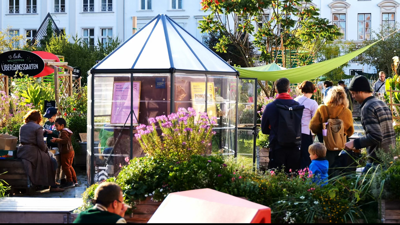 Ein lebendiger Gemeinschaftsgarten mit Menschen, die sich zwischen Pflanzen, Hochbeeten und einem kleinen Glashaus versammeln.